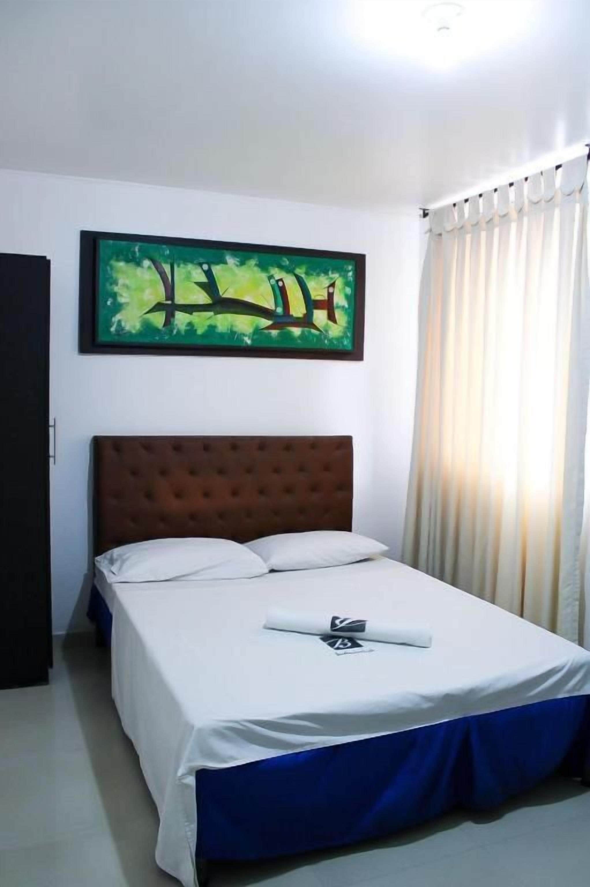 A hotel room with a bed, a headboard, a painting on the wall, and a window with curtains.