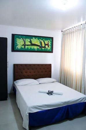 Una habitación de hotel con cama, cabecera, un cuadro en la pared y ventana con cortinas.