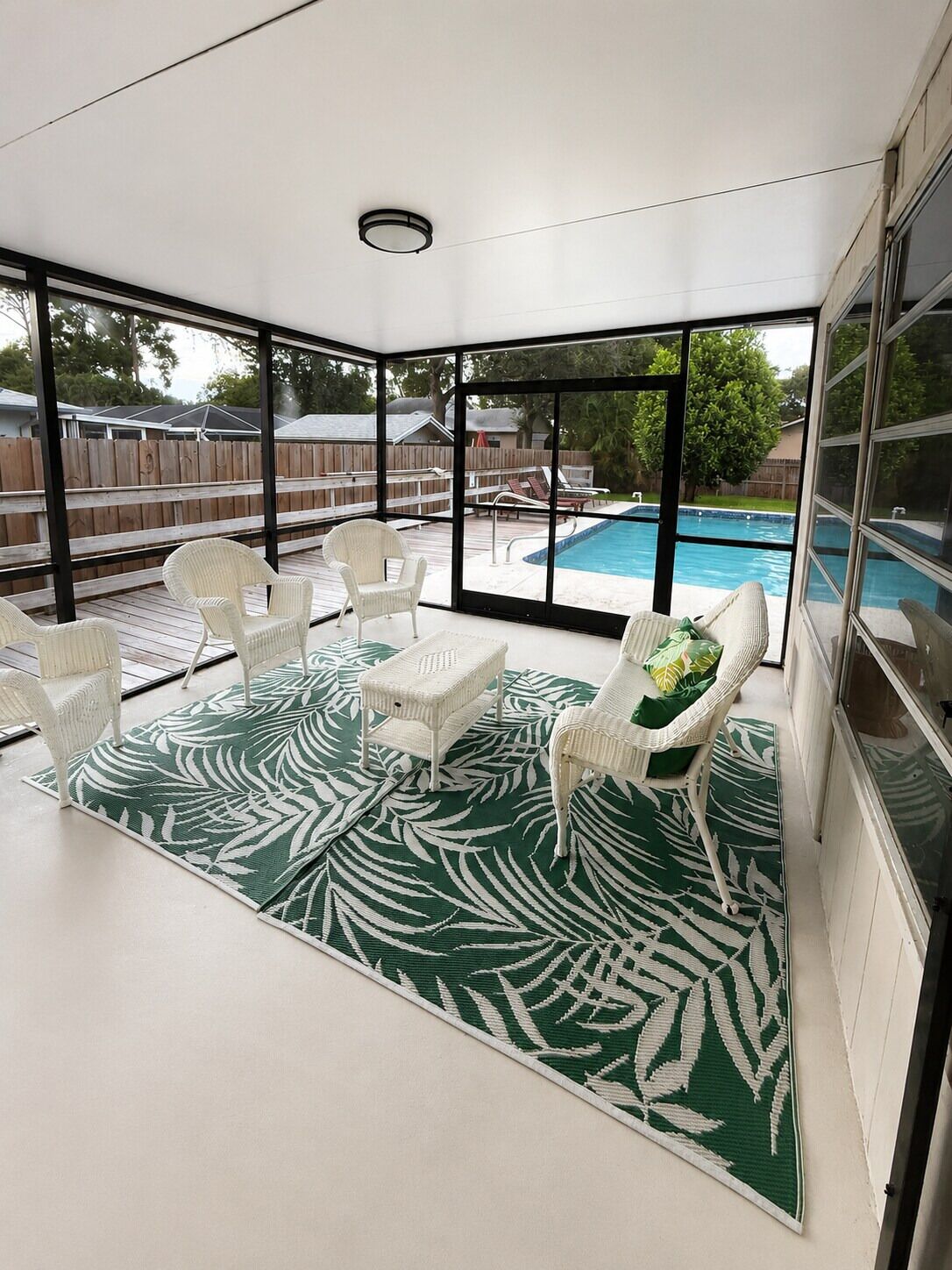A covered patio area with a pool, white wicker furniture, and a green patterned rug.