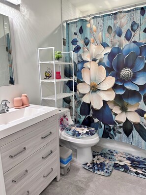 A bathroom with a white vanity, a toilet with a floral seat cover, a shower curtain with a floral pattern, and a white shelf unit with decorative items. - Professional Rental Suite (Penticton)