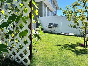 A well-maintained grassy yard with a white lattice fence, a small tree, and a building with a white exterior in the background. - Professional Rental Suite (Penticton)