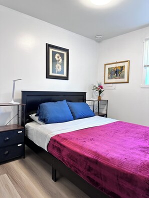 A bedroom with a bed, a nightstand, a bedside table, and two framed pictures on the wall. - Professional Rental Suite (Penticton)