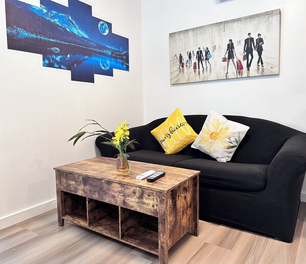 A living room with a black sofa, a wooden coffee table with a vase of yellow flowers, and artwork on the walls. - Professional Rental Suite (Penticton)
