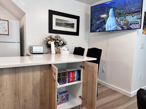 A modern room with a flat-screen TV mounted on the wall, a framed picture above the TV, a small table with a toaster, a vase with flowers, and a bookshelf with books. - Professional Rental Suite (Penticton)