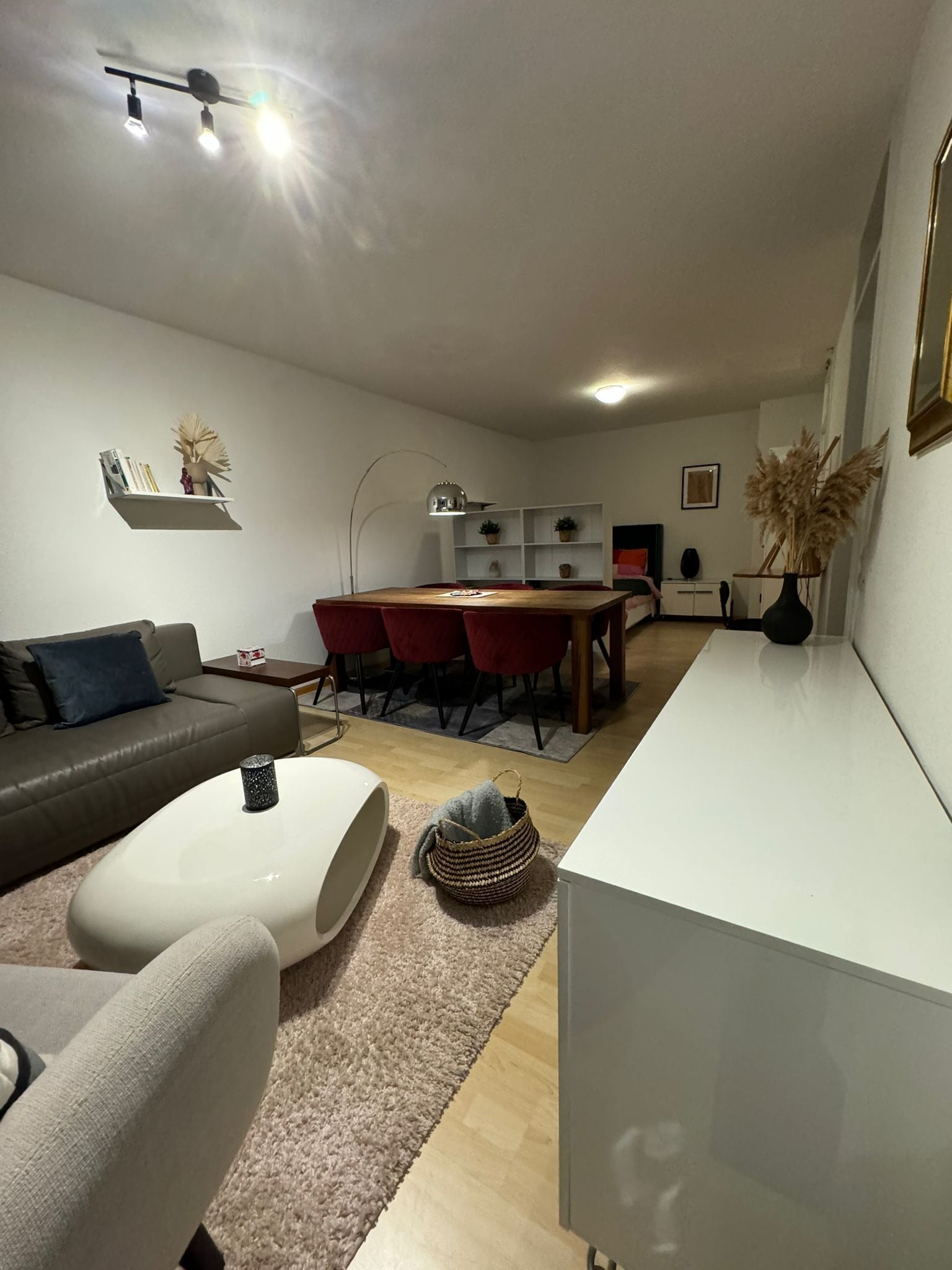 A modern living room with a sofa, a coffee table, a dining area with a table and chairs, and a kitchen area with cabinets and a sink.