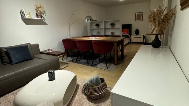 A modern living room with a sofa, a coffee table, a dining area with a table and chairs, and a kitchen area with cabinets and a sink. - Stylish flat close to Basel with a stunning view (Weil am Rhein)