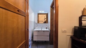 Un pasillo estrecho que conduce a un baño con lavabo y espejo blancos, puertas de madera y suelo de baldosas.