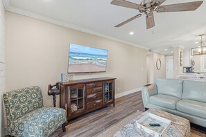 Un salon avec un ventilateur de plafond, un téléviseur fixé au mur, un meuble en bois, un fauteuil à motifs et un canapé bleu clair.