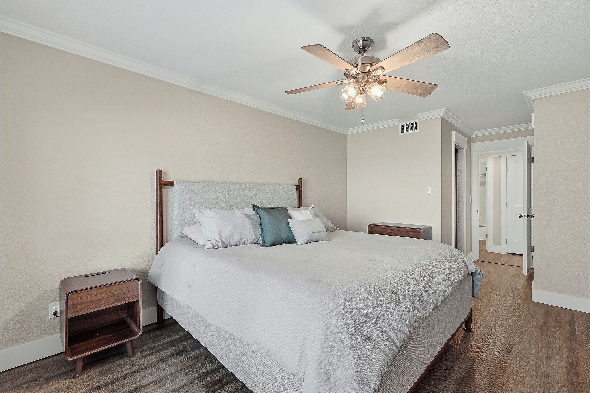 A bedroom with a bed, nightstands, a ceiling fan, and a door leading to another room.