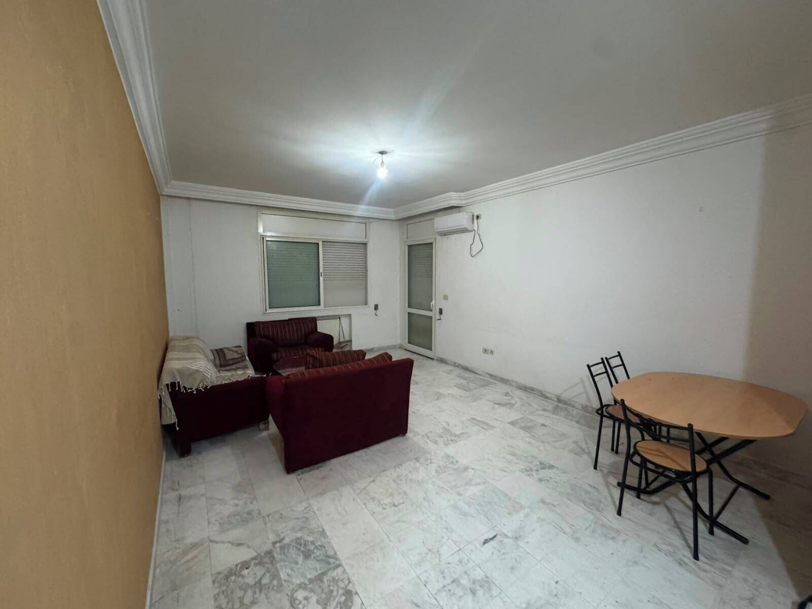 Ein Zimmer mit einer Sofagarnitur, einem Esstisch mit Stühlen, einem Fenster mit Jalousien und einer Deckenleuchte.