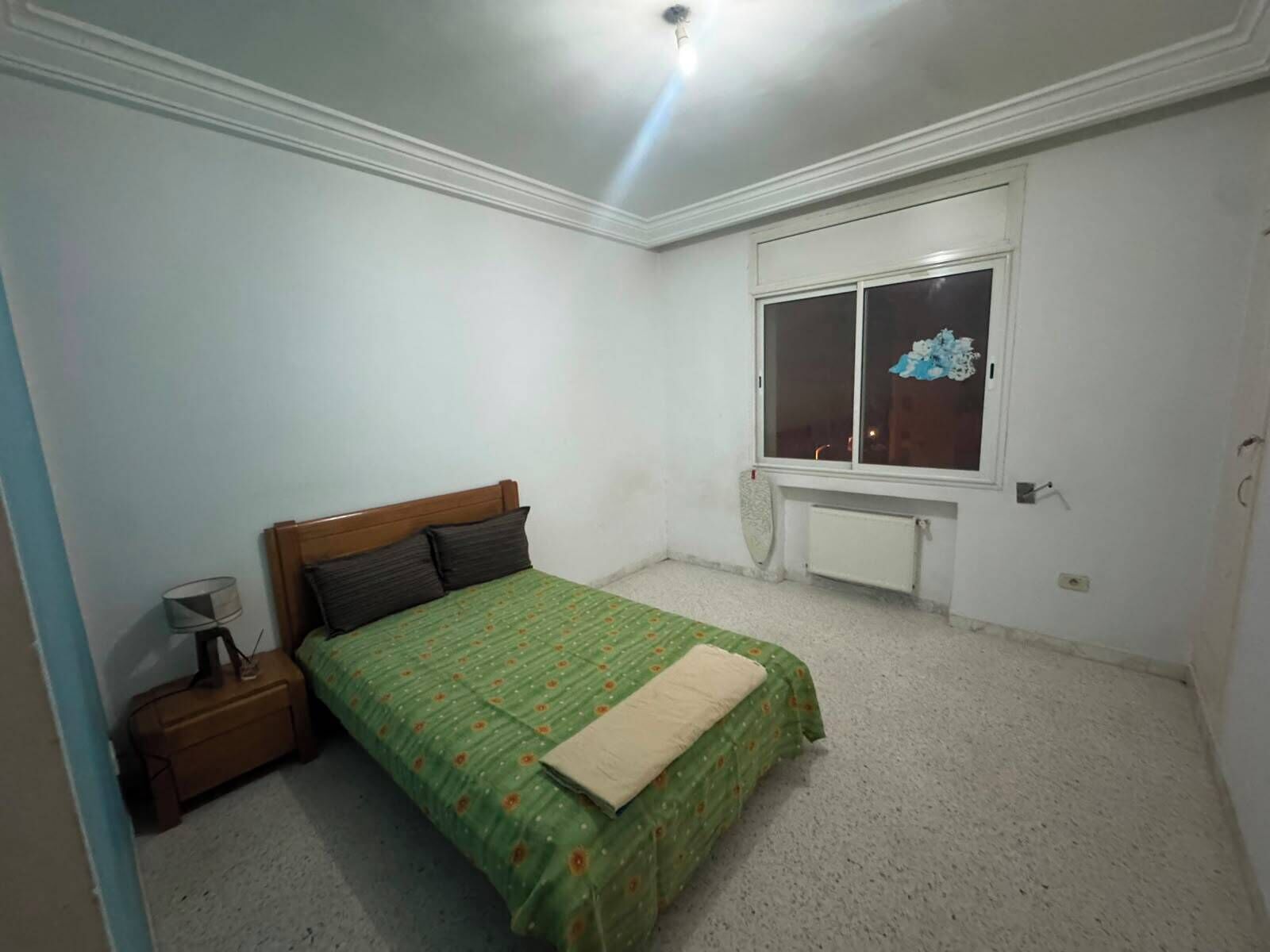 Une chambre avec un lit simple, une table de chevet, une fenêtre et un radiateur.