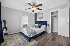 A bedroom with a bed, a ceiling fan, a window with blinds, a bedside table, and a door. - Blue Luxe Getaway Stylish 2BR Near Downtown (Houston)