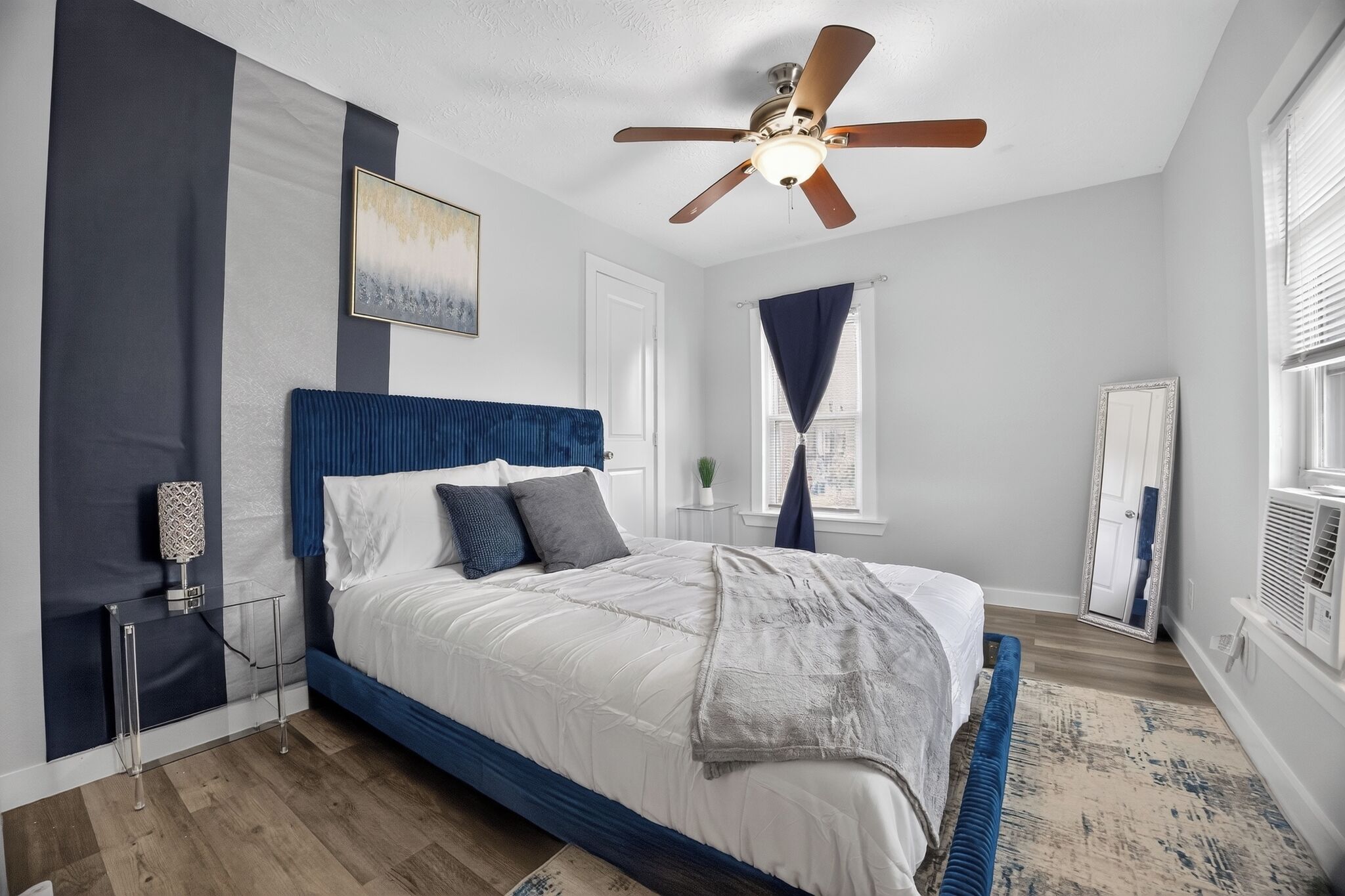 A bedroom with a bed, a ceiling fan, a window with curtains, a mirror, and a bedside table.