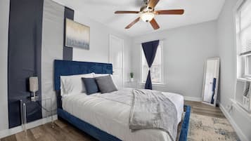 Un dormitorio con cama, ventilador de techo, ventana con cortinas, espejo y mesita de noche.