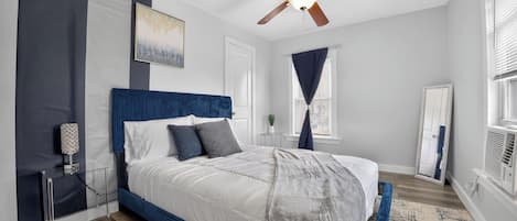 Une chambre à coucher avec un lit, un ventilateur de plafond, une fenêtre avec des rideaux, un miroir et une table de chevet.