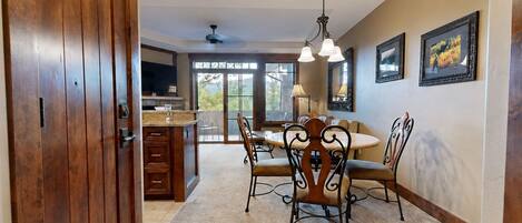 A dining area with a wooden table and chairs, a kitchen in the background, and large windows.