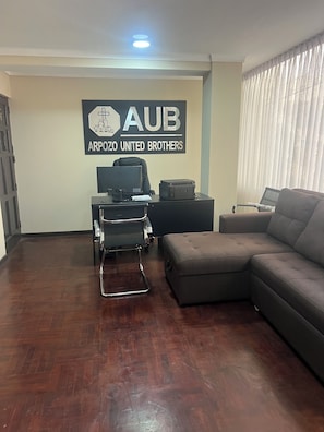 Una habitación con piso de madera oscura, una silla de oficina negra, un escritorio negro con una computadora, una impresora y un sofá gris grande. Un letrero en la pared dice 'AUB ARPOZO UNITED BROTHERS'.