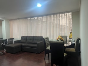 Una sala de estar con un sofá modular, una mesa de comedor con sillas y un ventanal con cortinas translúcidas.