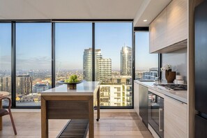A modern kitchen with a large window, a dining area, and a view of the cityscape. - Panoramic City View, Stunning 3BR Pool & Gym & Parking (South Yarra)