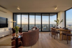 A modern living room with a curved sofa, a dining area with a wooden table and chairs, and a large window offering a city view. - Panoramic City View, Stunning 3BR Pool & Gym & Parking (South Yarra)