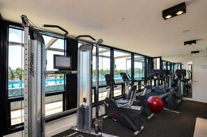 A gym with cardio machines, weightlifting equipment, and a large window offering a view of a pool. - Panoramic City View, Stunning 3BR Pool & Gym & Parking (South Yarra)