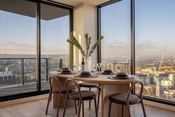 Un coin repas avec une table ronde dressée pour quatre personnes, de grandes fenêtres donnant sur la ville et un vase agrémenté de verdure placé sur la table.