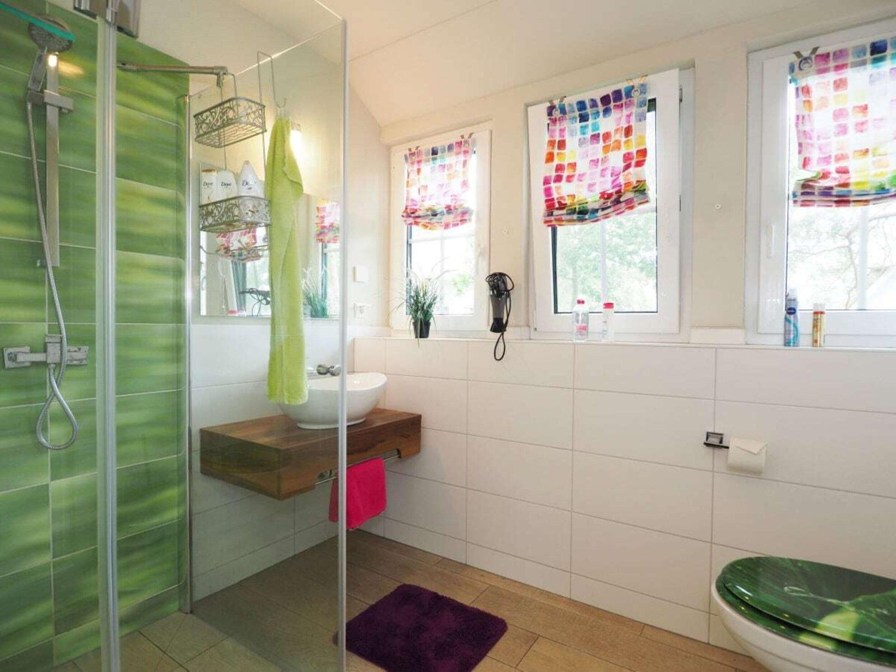 Un baño con cabina de ducha de vidrio, un lavamanos blanco, un mueble de tocador de madera, un inodoro verde y una alfombra púrpura.