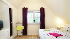 Quarto com cama, janela com cortinas, mesa de cabeceira com abajur e televisão em móvel.