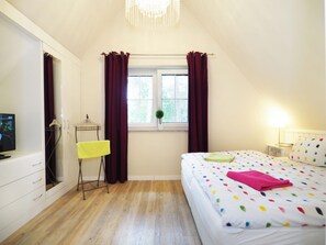 Ein Schlafzimmer mit einem Bett, einem Fenster mit Vorhängen, einem Nachttisch mit Lampe und einem Fernseher auf einem Schrank.