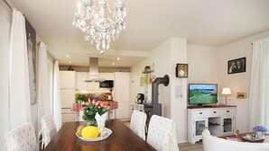 Uma sala de jantar com uma mesa de madeira, cadeiras, um lustre, uma cozinha ao fundo e uma televisão sobre um suporte.