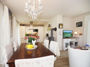 Ein Essbereich mit Holztisch, Stühlen, einer Chandelüster, einer Küche im Hintergrund und einem Fernseher auf einem Standfuß.
