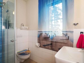 Ein Badezimmer mit Duschkabine aus Glas, einer Toilette mit farbigem Sitz, einem Waschbecken und einem Handtuchhalter mit einem roten Handtuch.