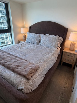 A bedroom with a large bed, two bedside tables, and a window with a view of buildings. - 3-bedroom Condo in Kelowna on Lake Okanagan with AC and Gym (Kelowna)