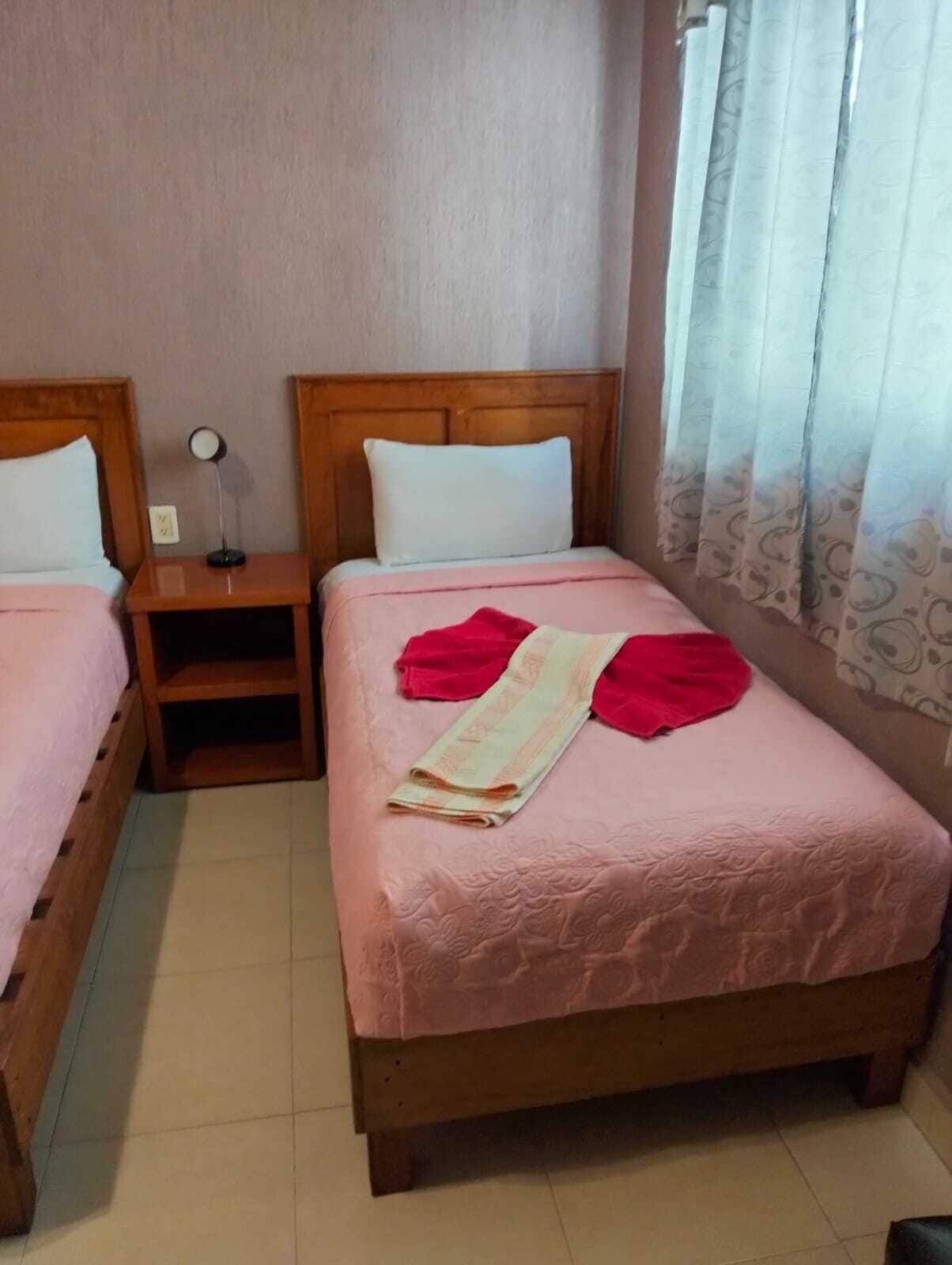 Habitació d'hotel amb dos llits individuals, una taula de nit amb una làmpada i una finestra amb cortines.
