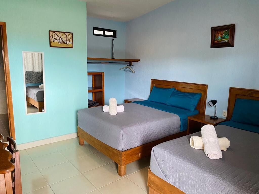 Una habitación con dos camas de madera, cada una con cubrecama gris y almohadas azules, y una pequeña mesa con una silla.