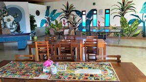 Un comedor con mesas y sillas de madera, una mesa de billar y un mural con plantas tropicales en la pared.