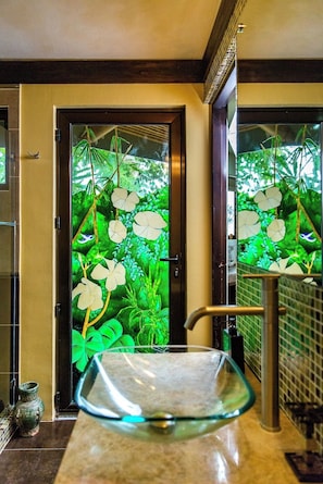 Un baño con un lavamanos de vidrio, una puerta con un diseño de hojas verdes y un piso de baldosas.