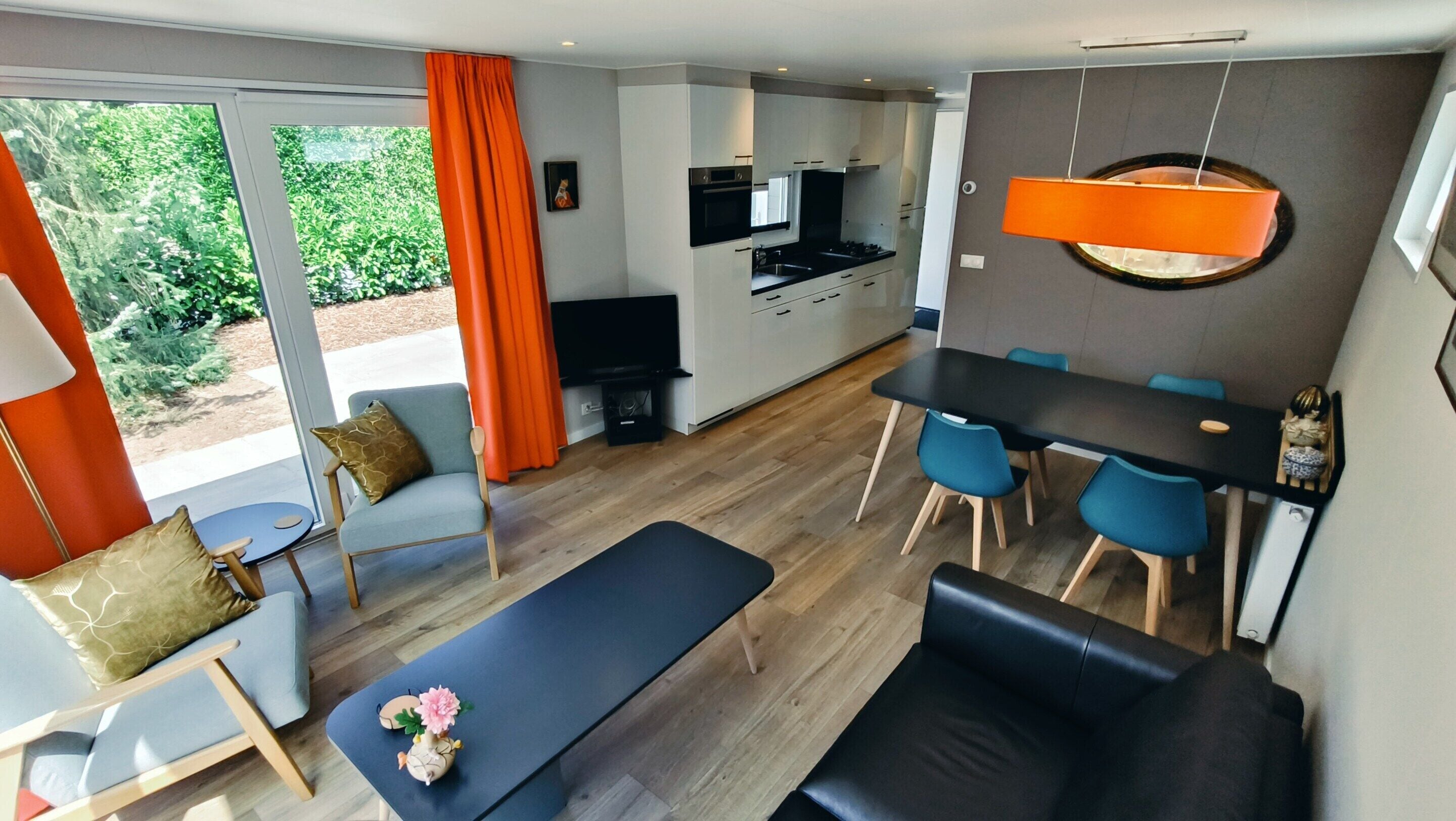A modern living room with a sofa, a dining table with chairs, a kitchen area, and a large window with curtains.