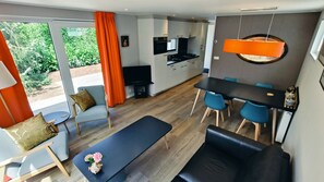 A modern living room with a sofa, a dining table with chairs, a kitchen area, and a large window with curtains. - Chalet Vogelzang for Retreat (Vorden)