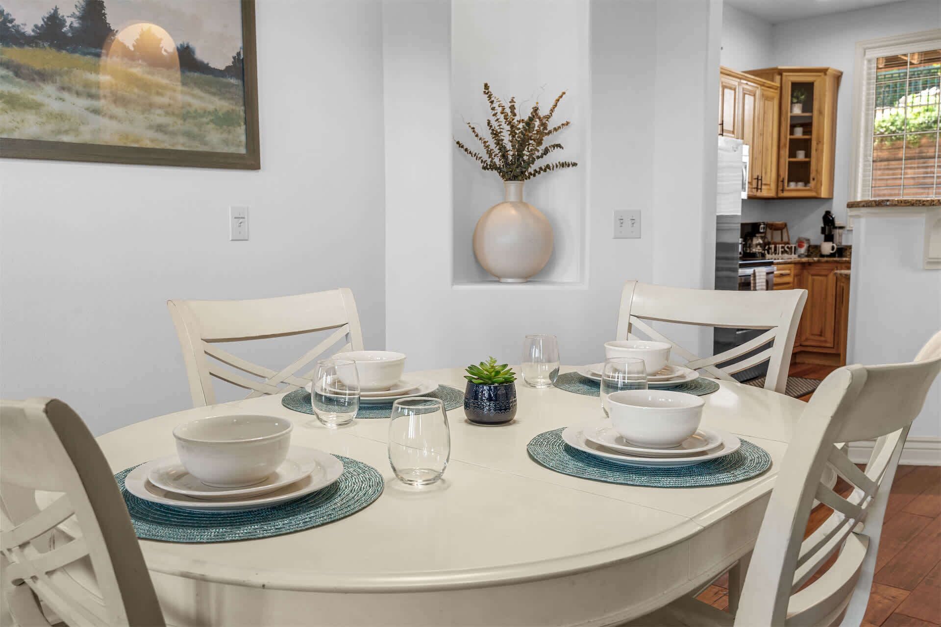 A dining area with a round table set for four, surrounded by white chairs. A vase with dried plants is placed on the wall above the table.
