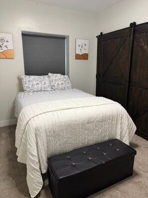 A bedroom with a bed, a window with blinds, a dark wooden wardrobe, and two wall-mounted artworks.