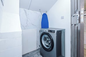 A compact laundry room with a washing machine, a blue drying rack, and a door with a handle. - 2 Bed | 7 Min to City Centre | Free Parking (Manchester)
