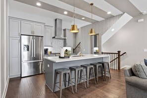 A modern kitchen with a stainless steel refrigerator, a central island with bar stools, and a staircase leading to an upper level. - Luxury 8BR Nashville Stay • 2 Rooftops + City Views + Fire Pits (Nashville)