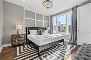 A bedroom with a bed, a nightstand, a lamp, a chandelier, and a window with curtains. - Luxury 8BR Nashville Stay • 2 Rooftops + City Views + Fire Pits (Nashville)