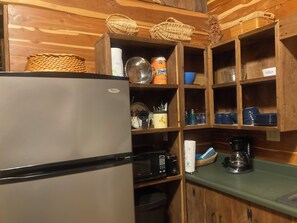 A compact kitchen with a refrigerator, microwave, and coffee maker. Wooden shelves hold various kitchen items and utensils. - Adorable 1-bedroom cabin in Treat, Arkansas.  2 adults, no pets, 2 night minimum (Dover)