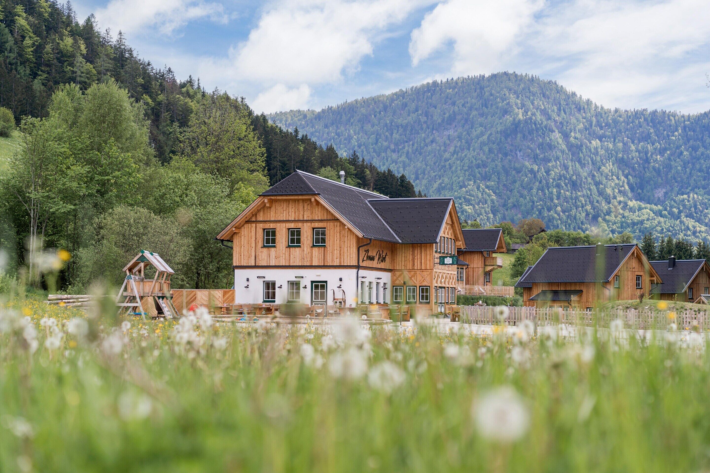 Ein holzgetägeltes Chalet mit einem geneigten Dach, eingebettet in eine üppige grüne Landschaft und mit einem bewaldeten Berg im Hintergrund.