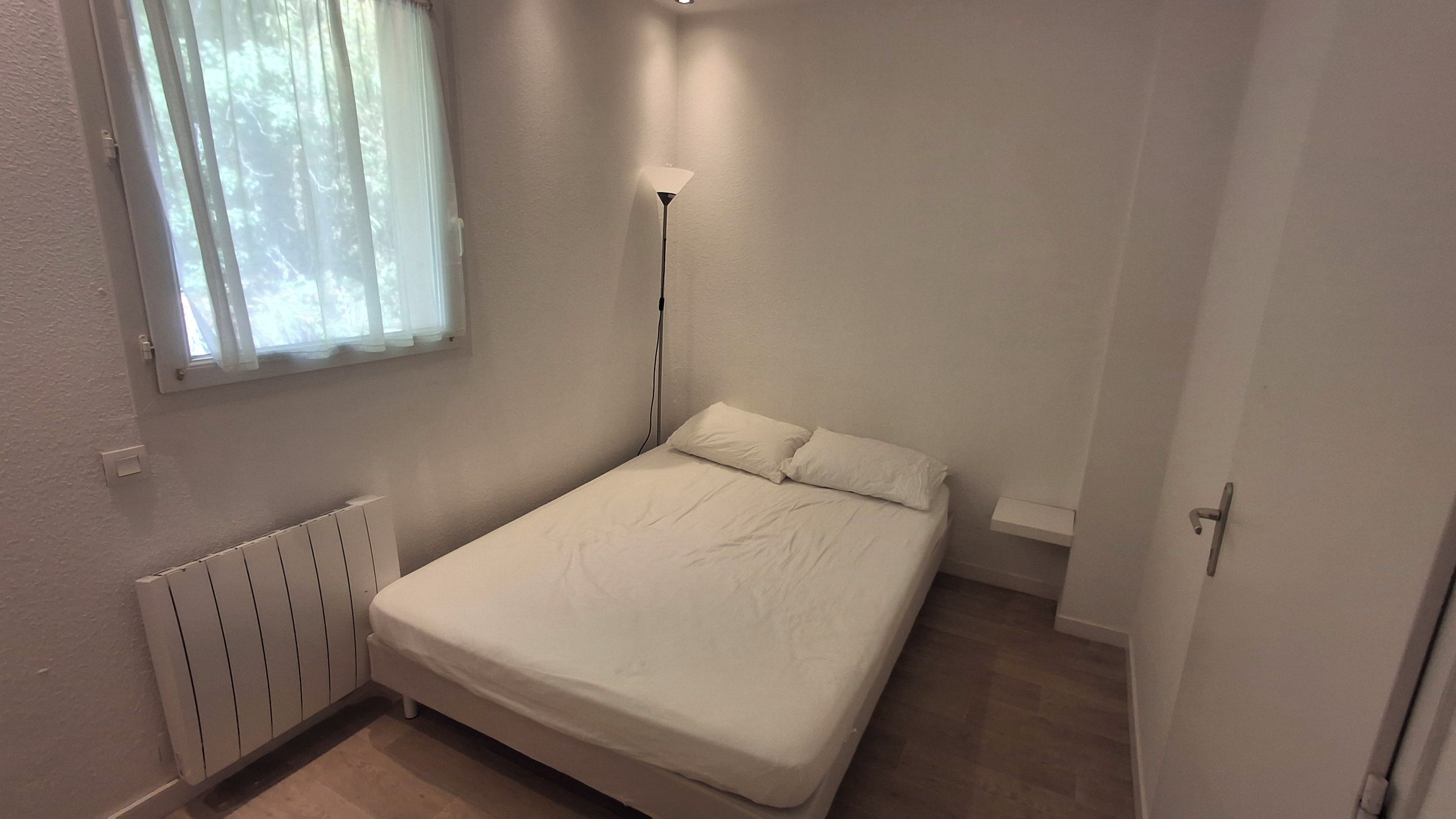 Una habitación pequeña con cama individual, colchón blanco, lámpara de noche, ventana con cortinas traslúcidas y un radiador.