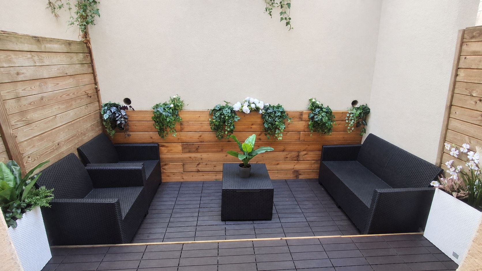 Een terras met zwart rieten meubilair en een houten wand met groene planten.