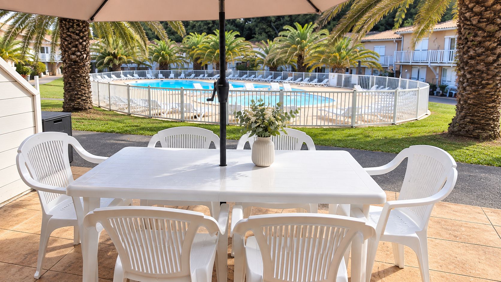 Un patio con una mesa y sillas, una piscina al fondo y una cerca blanca delimitando el espacio.