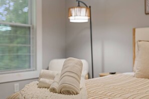 Una cama bien hecha con toallas enrolladas, una lámpara de noche y una ventana con persianas.
